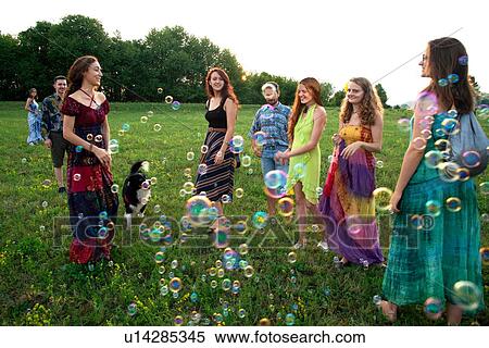 Stock Photography - Large group of happy young adults standing in field with floating bubbles. Fotosearch