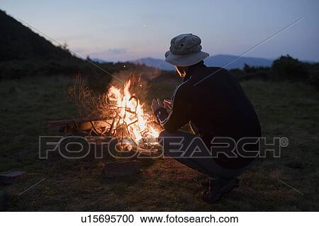 Stock Image - Man crouching by campfire. Fotosearch