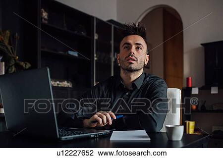 Stock Image - Man looking up while using laptop in home office. Fotosearch