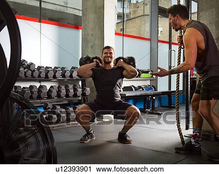 Stock Image - Man motivating friend doing squats, lifting kettlebells in gym. Fotosearch