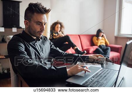 Man using laptop, friends relaxing in background in home office View Large Photo Image Stock Image - Man using laptop, friends relaxing in background in home office. Fotosearch