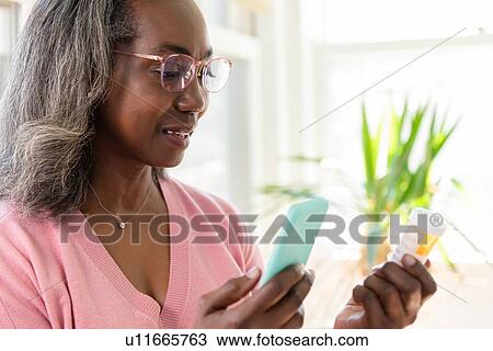 Stock Image - Mature woman holding smart phone and medicine bottle. Fotosearch