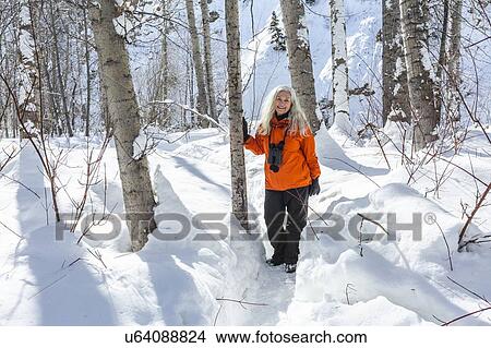 Picture - Mature woman wearing orange coat by bare trees in snow. Fotosearch
