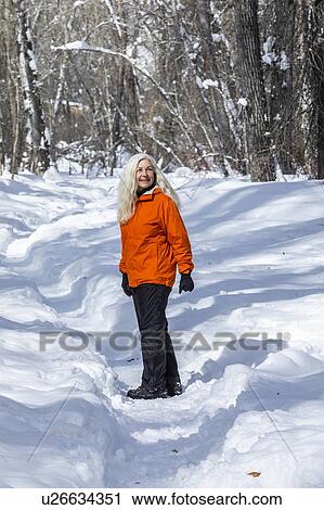 Stock Image - Mature woman wearing orange coat by bare trees in snow. Fotosearch