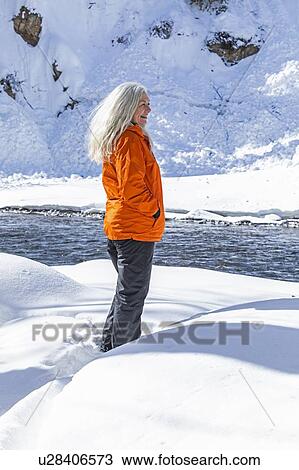 Stock Image - Mature woman wearing orange coat on snow by river. Fotosearch