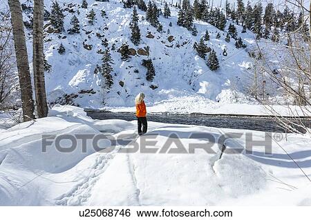 Stock Photograph - Mature woman wearing orange coat on snow by river. Fotosearch