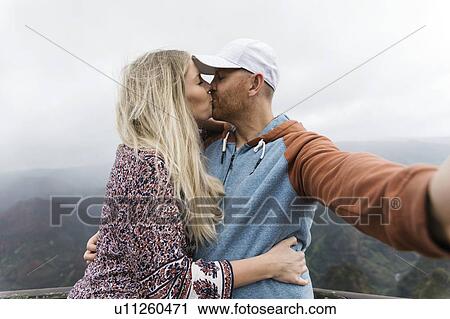Stock Image - Mid adult couple taking selfie while kissing. Fotosearch