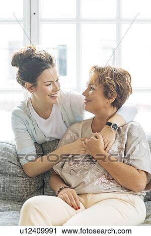Stock Image - Mother and adult daughter hugging on sofa. Fotosearch