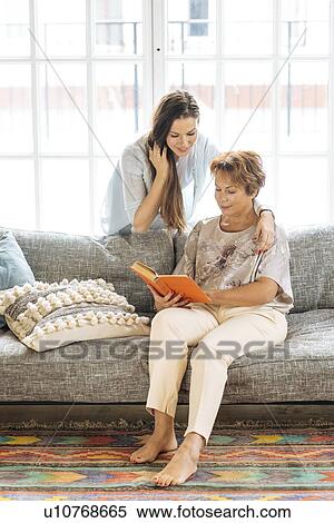 Stock Photography - Mother and adult daughter looking at book on sofa. Fotosearch