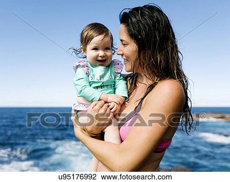 Stock Image - Mother carrying her baby girl wearing swimwear. Fotosearch