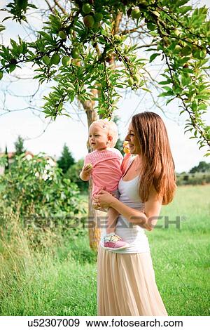 Stock Photo - Mother carrying toddler daughter under fruit tree, Arezzo, Tuscany, Italy. Fotosearch
