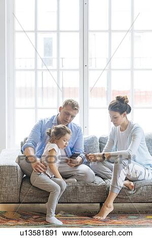 Parents holding smart phones on sofa with their daughter View Large Photo Image Stock Image - Parents holding smart phones on sofa with their daughter. Fotosearch