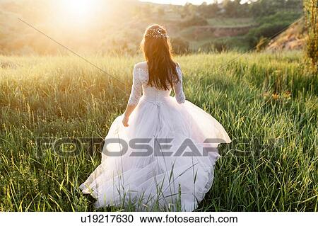 Stock Image - Rear view of bride in wedding dress walking in grass. Fotosearch
