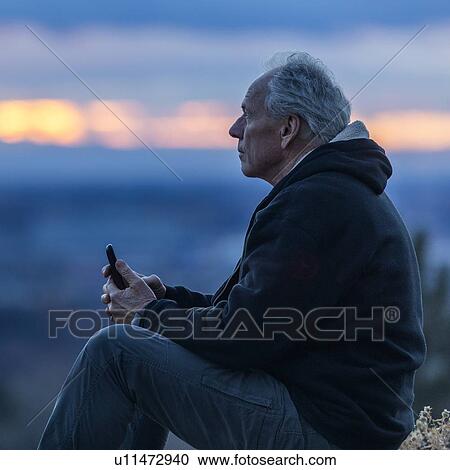 Stock Image - Seated man holding smart phone at sunset. Fotosearch