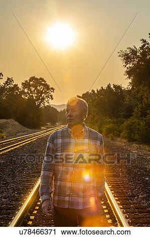 Stock Image - Senior man walking on railroad tracks at sunset. Fotosearch