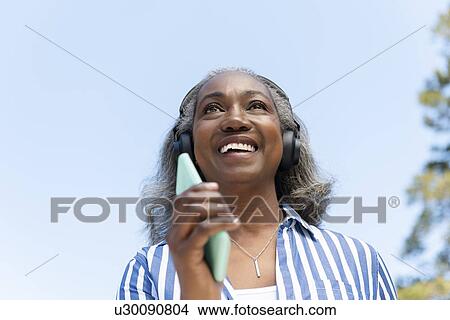 Smiling mature woman walking with headphones and smartphone View Large Photo Image Picture - Smiling mature woman walking with headphones and smartphone. Fotosearch