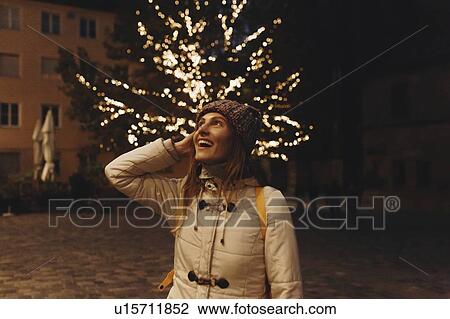 Stock Image - Smiling woman wearing coat by fairy lights in tree at night. Fotosearch