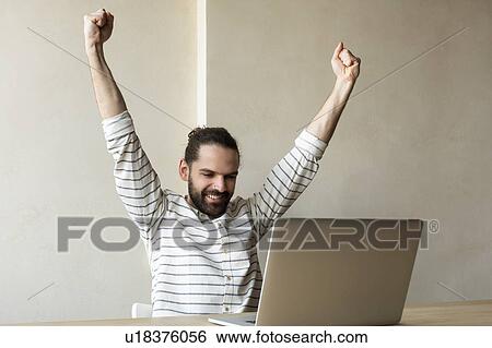Smiling young businessman with his arms raised by laptop View Large Photo Image Stock Photograph - Smiling young businessman with his arms raised by laptop. Fotosearch