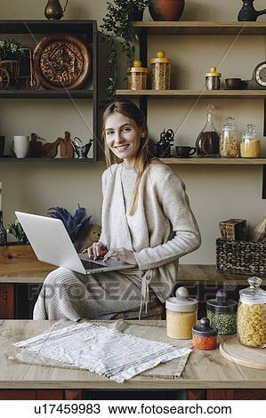 Stock Image - Smiling young woman using laptop in kitchen. Fotosearch