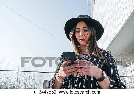 Stock Photo - Stylish young woman in felt hat outside office building looking at smartphone, Turin, Piemonte, Italy. Fotosearch