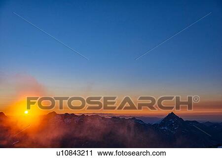 Stock Image - Sun glowing on horizon over mountain range, Saas-Fee, Valais, Switzerland. Fotosearch