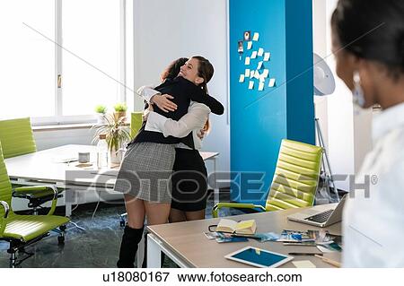 Stock Photo - Two female business creatives hugging in office, over shoulder view. Fotosearch
