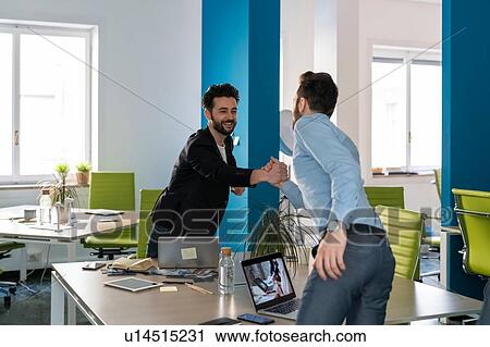 Stock Image - Two male business creatives smiling and shaking hands over office desk. Fotosearch