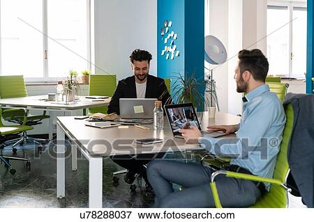 Stock Photo - Two male business creatives using laptops at office desk. Fotosearch