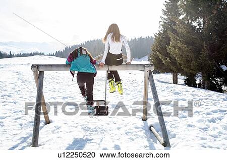 Stock Image - Two teenage girl skiers sitting on top of swing in snow covered landscape, rear view, Tyrol, Styria, Austria. Fotosearch