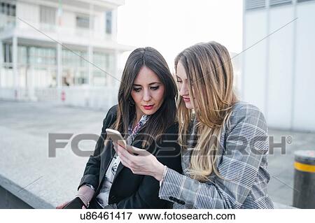 Stock Photo - Two young businesswomen sitting outside office building looking at smartphones, Turin, Piemonte, Italy. Fotosearch