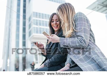 Stock Photo - Two young businesswomen sitting outside office building looking at smartphones, Turin, Piemonte, Italy. Fotosearch