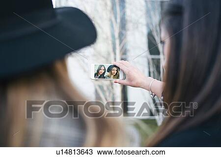Stock Image - Two young women taking smartphone selfie in city, over shoulder view. Fotosearch