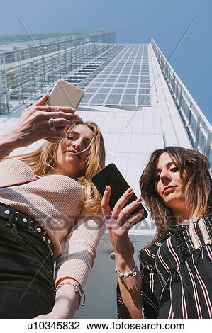 Stock Image - Two young women with smartphones in front of skyscraper, low angle portrait, Turin, Piemonte, Italy. Fotosearch