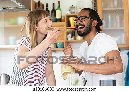 Stock Image - Woman feeding man with wooden spoon in kitchen. Fotosearch
