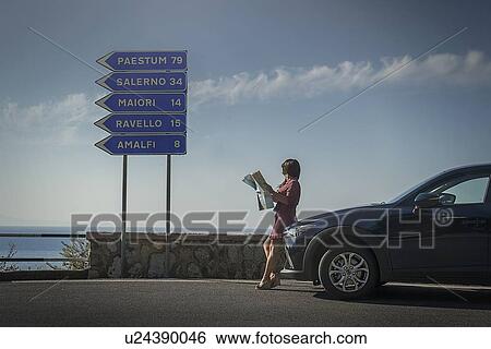 Stock Photograph - Woman holding map leaning on car by road sign. Fotosearch