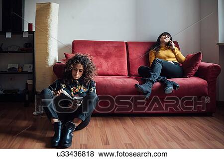Stock Photo - Woman reading book, friend talking on smartphone on sofa. Fotosearch