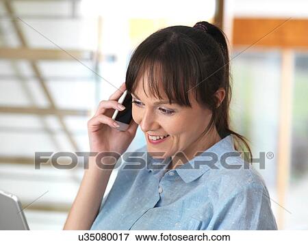 Stock Photo - Woman smiling over the phone during a conversation at her desk. Fotosearch