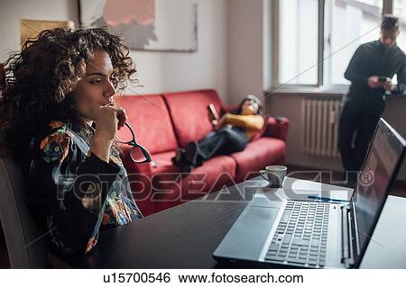 Stock Photograph - Woman using laptop, friends relaxing in background in home office. Fotosearch