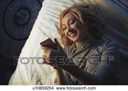 Woman using smart phone while lying on bed View Large Photo Image Picture - Woman using smart phone while lying on bed. Fotosearch