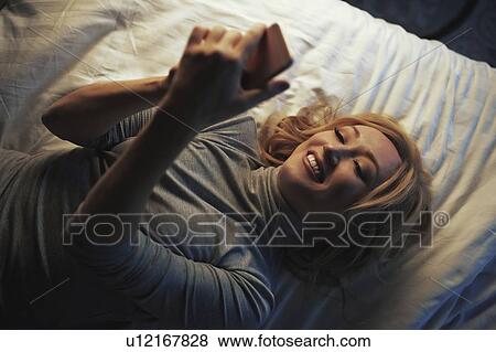 Stock Photo - Woman using smart phone while lying on bed. Fotosearch