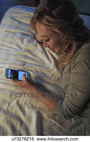 Stock Photograph - Woman using smart phone while lying on bed. Fotosearch