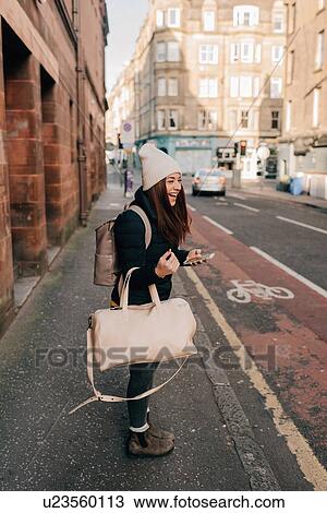 Stock Image - Woman waiting on kerb with cellphone and luggage, Edinburgh, Scotland. Fotosearch