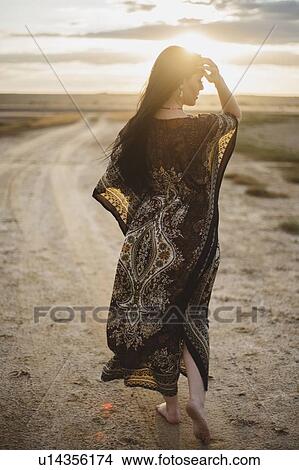 Woman wearing patterned dress walking on dirt road at sunset View Large Photo Image Picture - Woman wearing patterned dress walking on dirt road at sunset. Fotosearch