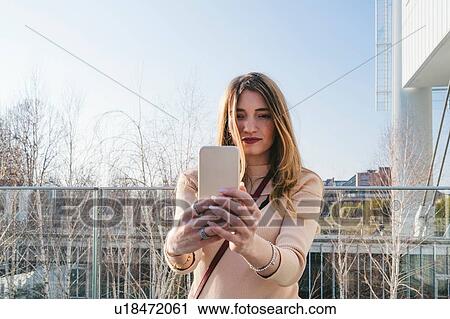 Stock Image - Young blond woman outside office building taking smartphone selfie, Turin, Piemonte, Italy. Fotosearch