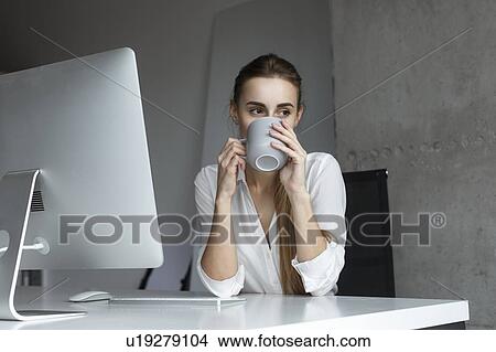 Picture - Young businesswoman drinking coffee at desk. Fotosearch