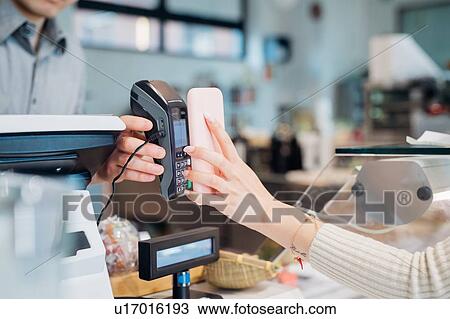 Stock Image - Young businesswoman making smartphone payment at cafe counter, cropped. Fotosearch