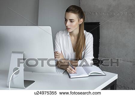 Young businesswoman taking notes while working at desktop computer View Large Photo Image Stock Photography - Young businesswoman taking notes while working at desktop computer. Fotosearch