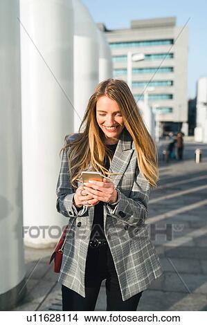 Picture - Young businesswoman with long blond hair looking at smartphone outside office building, Turin, Piemonte, Italy. Fotosearch