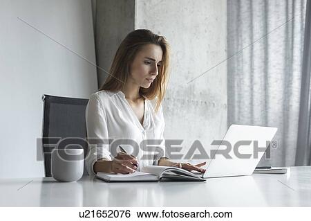 Stock Photograph - Young businesswoman writing notes while working on laptop. Fotosearch