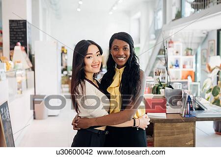 Stock Image - Young businesswomen hugging in bookshop, portrait. Fotosearch